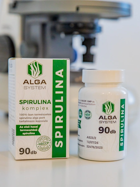 Alga System Spirulina Komplex kapszula és doboza laboratóriumi háttérrel – 90 darabos kiszerelés, 100%-ban természetes spirulina algaport tartalmaz, HACCP tanúsítvánnyal.