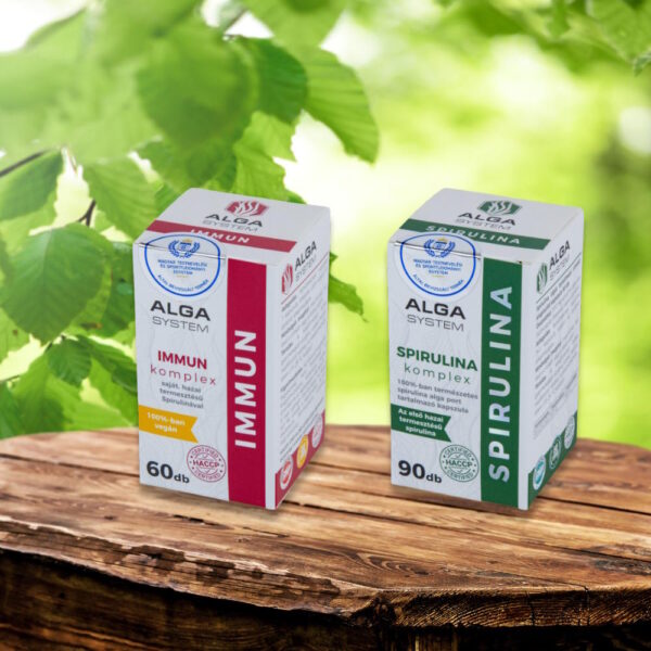Alga System Spirulina és Immun Komplex étrendkiegészítők dobozban, fa felületen, zöld lombos háttérrel.
