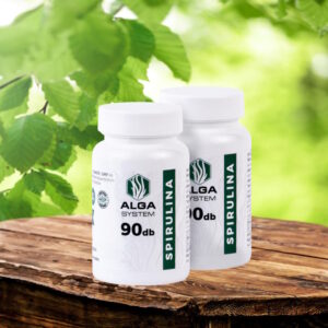 Alga System Spirulina Komplex 2x90 db-os, fehér, címkés flakonok, fa asztalon, zöld levelekkel a háttérben.